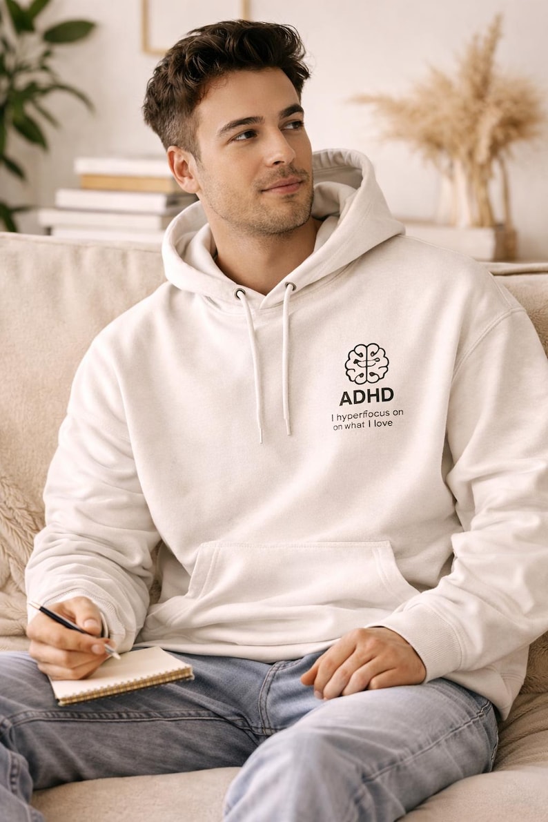 May include: Cream-colored hoodie with the text "ADHD" and a brain graphic. The text "I hyperfocus on what I love" is below the graphic. The hoodie has a front pocket and drawstring hood.
