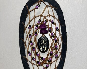 May include: A brown and black dreamcatcher with purple beads and a black and white skeleton charm hanging from it.