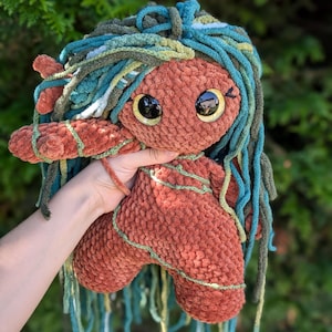 May include: A crocheted doll with long, teal, green, and white yarn hair. The doll has a brown body with a textured surface and large, black eyes with gold pupils.