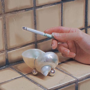 May include: A decorative seashell ashtray with a mother-of-pearl finish and three silver feet. A lit cigarette is held above the ashtray. The ashtray is on a tiled surface.