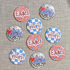 May include: Eight circular pinback buttons featuring various designs: red and white gingham with 'Eagles', blue and white checkerboard with 'Let's Go Eagles', and silver disco ball patterns with 'Arca Eagles' and a red heart graphic.