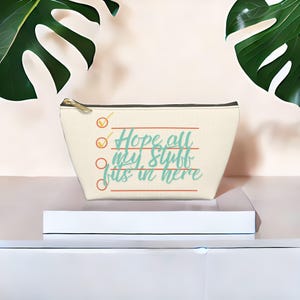 May include: Cream-colored cosmetic bag with a gold zipper and the text "Hope all my stuff fits in here" in teal script. The design includes orange check marks and lines. The bag is on a white platform with green leaves in the background.