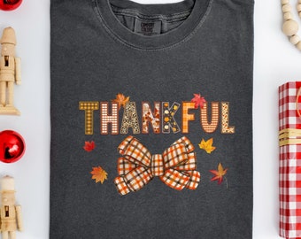 Könnte beinhalten: Ein dunkelgraues T-Shirt mit dem Wort "THANKFUL" in bunten gemusterten Buchstaben und einer großen karierten Schleife. Die Buchstaben und die Schleife sind von kleinen Herbstblättern umgeben.