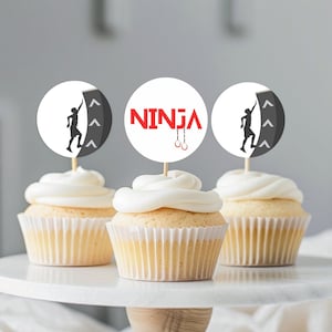 Puede incluir: Tres cupcakes blancos con glaseado blanco cubiertos con adornos redondos de papel para cupcakes. Los adornos representan la silueta de una persona escalando una pared con la palabra "NINJA" en letras rojas y un par de anillos de gimnasia rojos.