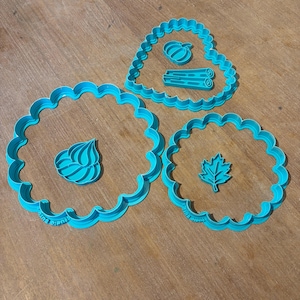 Op de afbeelding: Drie fel turquoise plastic koekjesvormpjes met geschulpte randen. Een grote ronde vorm bevat een toefje, een middelgrote hartvormige vorm bevat een pompoen en kaneelstokjes, en een kleine ronde vorm bevat een esdoornblad.