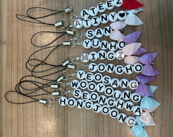 May include: A set of 8 phone charms featuring the names of the members of the K-pop group Ateez. Each charm is made of white beads with black lettering and a small bow. The bows are in various colors, including red, white, pink, purple, and blue.