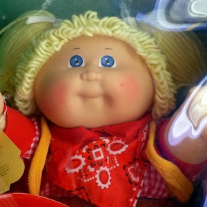 May include: A doll with blonde yarn hair styled in pigtails, blue eyes, and rosy cheeks. The doll wears a red and white checkered shirt, a red bandana with a white geometric pattern, and a yellow vest. A red plastic hat is visible in the foreground.