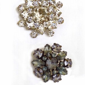 May include: Two circular brooches featuring faceted clear and grey crystals. The top brooch has a large central stone surrounded by square and round crystals. The bottom brooch features a central stone with marquise and round grey crystals.