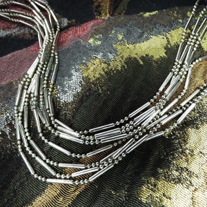 May include: A multi-strand necklace featuring silver-toned metallic beads and elongated cylindrical tube spacers. The design consists of several layered chains that create a cascading effect, showcasing a polished, reflective finish throughout the piece.