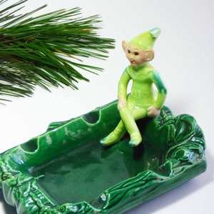 May include: Green ceramic ashtray shaped like a log with a green elf figurine sitting on top. The elf is wearing a green hat and has a green and white outfit.