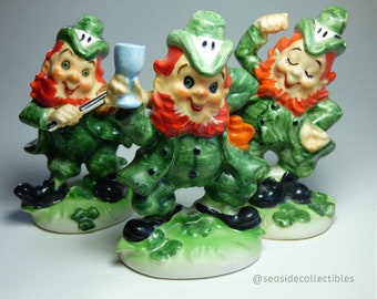 Seated Baby Green Elf Pixie Leprechaun Figurine Holding Shamrocks Clovers Peapod Green Suit Flower Hat Brown Hair Japan 1970s