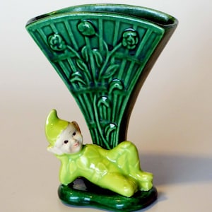 Op de afbeelding: Een groene keramische vaas met een fantasierijk ontwerp met een liggend elfje. De vaas heeft een waaiervormige opening en een groene en witte glazuur.