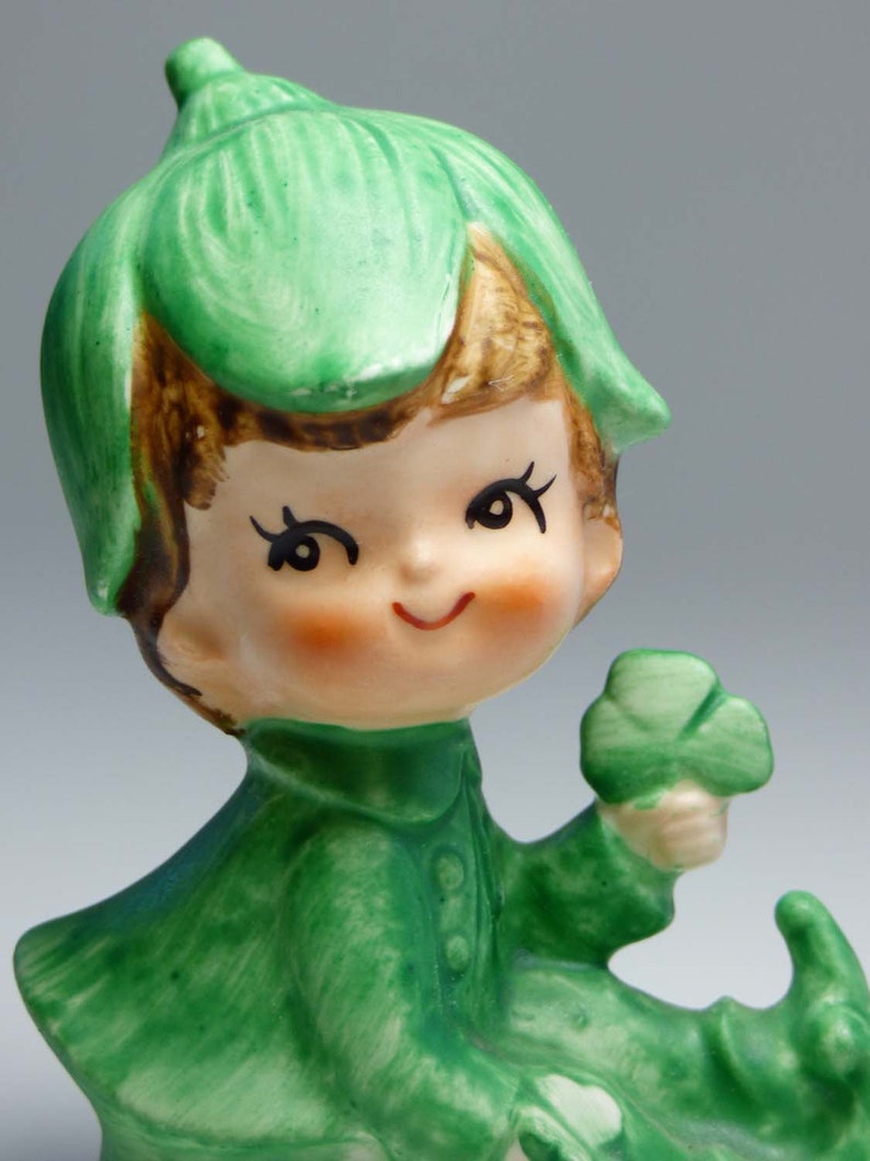 Seated Baby Green Elf Pixie Leprechaun Figurine Holding Shamrocks Clovers Peapod Green Suit Flower Hat Brown Hair Japan 1970s