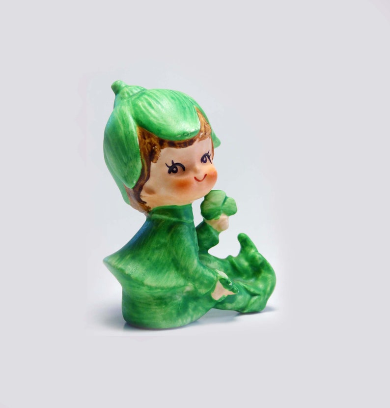 Seated Baby Green Elf Pixie Leprechaun Figurine Holding Shamrocks Clovers Peapod Green Suit Flower Hat Brown Hair Japan 1970s