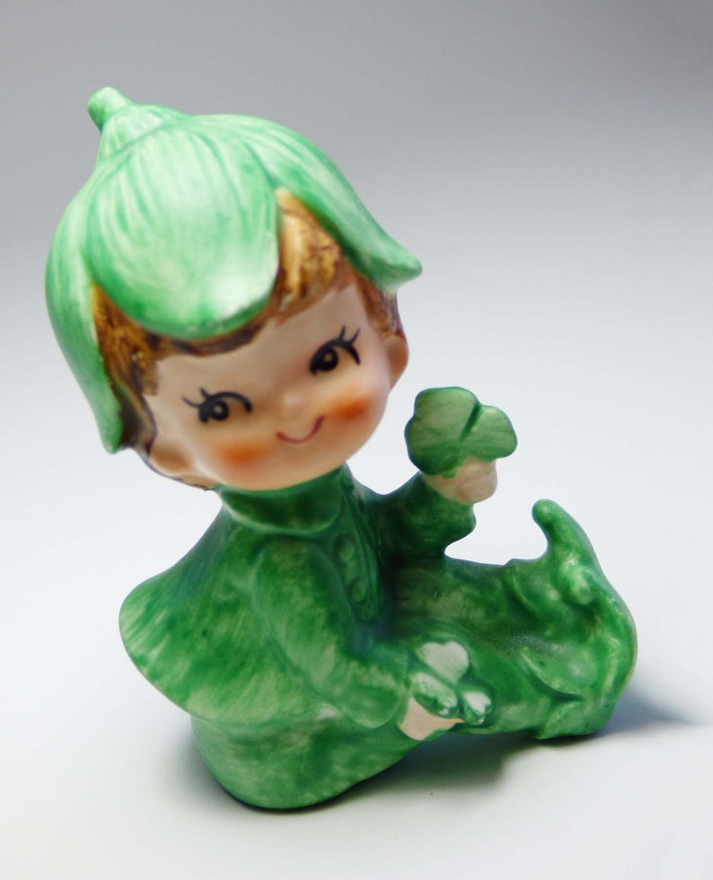 Seated Baby Green Elf Pixie Leprechaun Figurine Holding Shamrocks Clovers Peapod Green Suit Flower Hat Brown Hair Japan 1970s