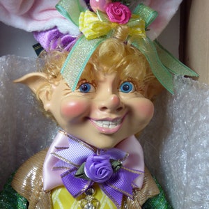 Könnte beinhalten: Eine fantasievolle Oster-Elfenpuppe mit blonden Haaren, die einen lila und grünen Hut mit einer rosa und gelben Schleife, eine lila und rosa Fliege und ein gelbes und grünes gemustertes Hemd trägt.