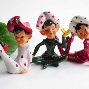 May include: Three vintage ceramic elf figurines. One wears a white outfit with red polka dots and a red hat. Another is in a green outfit, and the third is in red. Each has a white hat with polka dots.