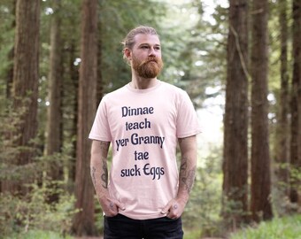 May include: A light pink t-shirt with the text "Dinnae teach yer Granny tae suck Eggs" in black. The shirt is worn by a person standing in a forest setting, wearing black pants.
