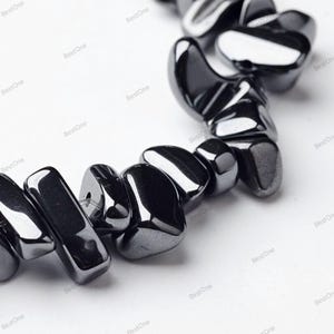 Può includere: Una collana di perline di ematite nera lucidata. Le perline sono di forma irregolare, con una superficie liscia e riflettente. Le perline sono infilate insieme, creando una collana o un braccialetto.