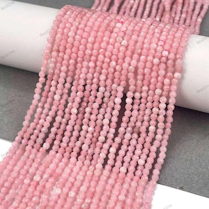 Può includere: Un filo di perline di pietre preziose rosa. Le perline sono sfaccettate e hanno una forma leggermente irregolare. Le perline sono infilate su un filo trasparente.