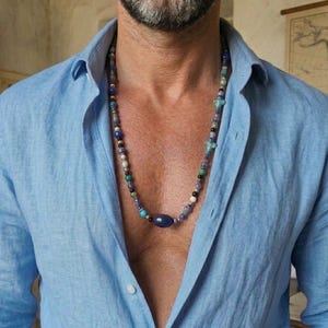 Può includere: Una collana di perline con pietre blu, verdi e bianche. La collana presenta una grande pietra blu a forma ovale come punto focale. Indossata sopra una camicia azzurra abbottonata.