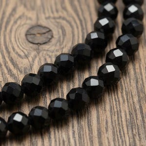 May include: Close-up of two strands of faceted black beads. The beads have a geometric, multi-faceted design, reflecting light. The background is a textured, wooden surface, providing a contrast to the smooth, dark beads. The beads are likely for jewelry making.