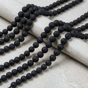 May include: Multiple strands of matte black beads are displayed against a light grey backdrop. The spherical beads are consistent in size, offering a dark, textured contrast. They appear to be strung together, potentially for crafting jewellery. The beads are likely measured in centimetres.