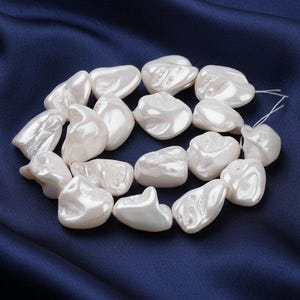 Op de afbeelding: Een streng onregelmatige, witte parels met een glanzend oppervlak. De parels zijn geregen op een witte draad. De achtergrond is een donkerblauwe, zijdeachtige stof. De parels hebben een unieke, organische vorm.