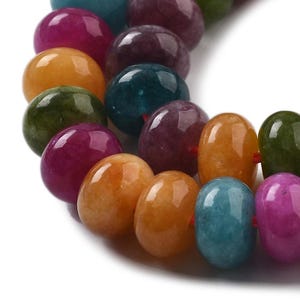 Può includere: Primo piano di una collana di perline di pietre preziose colorate. Le perline sono un mix di forme rotonde e ovali nelle tonalità del viola, arancione, verde e verde acqua. Le perline sono infilate con un filo rosso.