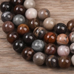 Può includere: Una collezione di perline di pietre preziose lucide in vari colori, tra cui marrone, grigio e crema. Le perline sferiche hanno una finitura lucida e sono disposte su una superficie di legno. Le perline variano per dimensioni e motivo.