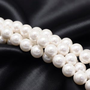 Può includere: Primo piano di una collana di perle bianche e lucenti. Le perle sono perfettamente rotonde e di dimensioni uniformi, appoggiate su un tessuto di raso nero. L'illuminazione mette in risalto la superficie liscia e riflettente delle perle.
