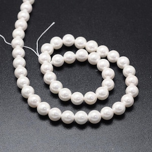 Op de afbeelding: Een streng witte, ronde parels, gepresenteerd tegen een donkere achtergrond. De parels zijn uniform van grootte en hebben een glad, glanzend oppervlak. Het draad is zichtbaar, wat suggereert dat de parels bedoeld zijn voor sieraden of presentaties.