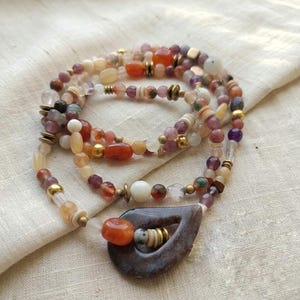Può includere: Una collana a più fili con una varietà di perline colorate nelle tonalità dell'arancione, del viola, del bianco e dell'oro. La collana include un grande ciondolo a forma di goccia con un ritaglio centrale e una grande perla arancione.