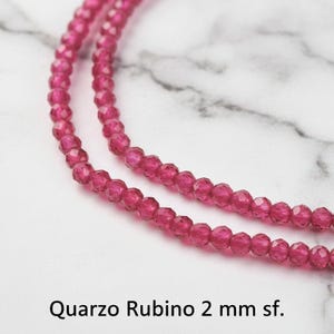 Può includere: Primo piano di due fili di perline di quarzo rosa scuro sfaccettate. Le perline sono infilate insieme, creando un aspetto simile a una collana. Il testo "Quarzo Rubino 2 mm sf." è in basso nell'immagine.