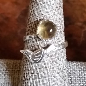 Könnte beinhalten: Ein silberner Ring mit einem kleinen Vogeldesign und einem runden, gelben Edelstein.