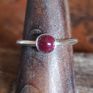 May include: A silver ring featuring a round, deep red gemstone. The gemstone is set in a bezel setting, and the band is a simple, polished silver. The ring is displayed on a wooden stand, showcasing its design.