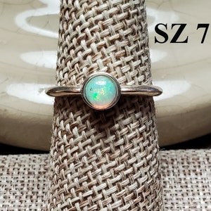 Puede incluir: Un anillo de plata con una piedra redonda de ópalo iridiscente. El anillo está en un expositor y el texto "SZ 7" es visible en la imagen.