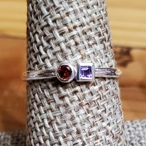 Puede incluir: Un anillo de plata con una piedra preciosa redonda de color naranja y una piedra preciosa cuadrada de color morado.