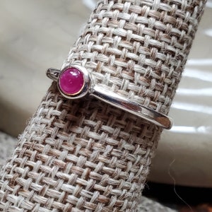 May include: A silver ring with a pink gemstone set in a bezel setting. The ring is on a fabric ring display stand.