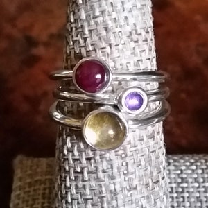 May include: Three silver rings with colorful gemstones. The top ring has a red gemstone, the middle ring has a purple gemstone, and the bottom ring has a yellow gemstone.