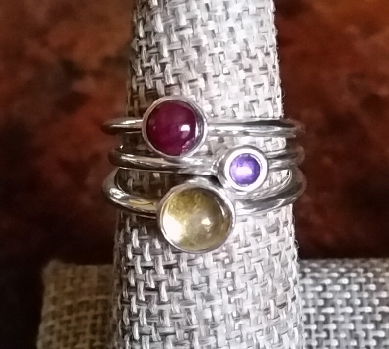 May include: Three silver rings with colorful gemstones. The top ring has a red gemstone, the middle ring has a purple gemstone, and the bottom ring has a yellow gemstone.