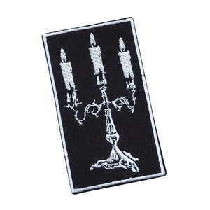 Puede incluir: Parche bordado negro con un diseño de candelabro blanco con tres velas. El candelabro tiene un diseño decorativo y ornamentado.
