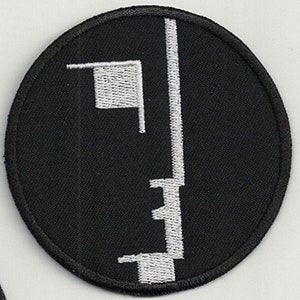 Puede incluir: Parche bordado negro con un diseño de cara estilizado blanco. La cara está compuesta por una línea vertical con una pequeña forma cuadrada en el lado izquierdo.