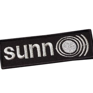 Puede incluir: Parche bordado negro con letras blancas que dicen "sunno" y un círculo blanco con círculos concéntricos a su alrededor.