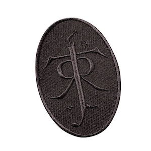 Puede incluir: Un parche bordado marrón oscuro en forma ovalada. El parche presenta un símbolo estilizado con una línea vertical y elementos curvos, posiblemente un logotipo o emblema. El fondo es de tela texturizada.