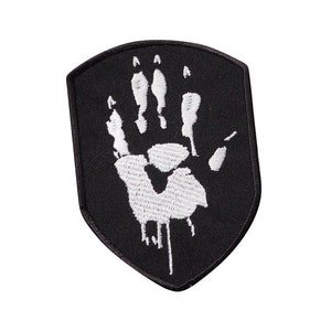 May include: Black embroidered patch featuring a white handprint in the centre. The handprint is stylised and has five fingers. The patch is shaped like a shield.