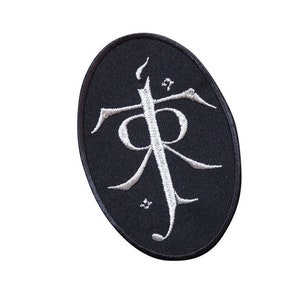 Può includere: Una toppa ricamata nera con un simbolo bianco, che ricorda una lettera stilizzata "R" con un design a forma di fiamma sopra. Il simbolo è circondato da piccole stelle bianche.