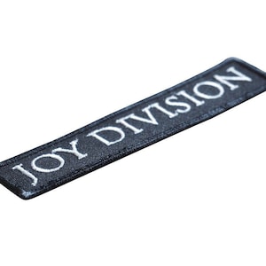 Pode incluir: Um remendo de tecido preto com texto bordado branco que diz "JOY DIVISION".