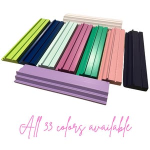 May include: A collection of rectangular wooden display stands featuring a grooved channel along the top surface. The stands are finished in various solid colors including lime green, navy blue, mint, purple, pink, and white. Text reads All 33 colors available.