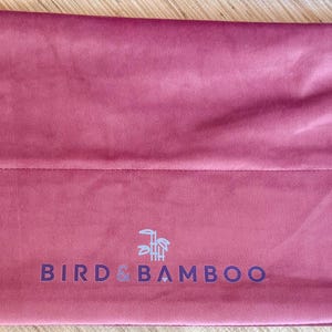 May include: A dusty rose-colored velvet bag with a drawstring closure. The bag features the text "BIRD & BAMBOO" in a dark blue font. The drawstring is white with wooden beads. The bag is rectangular in shape.